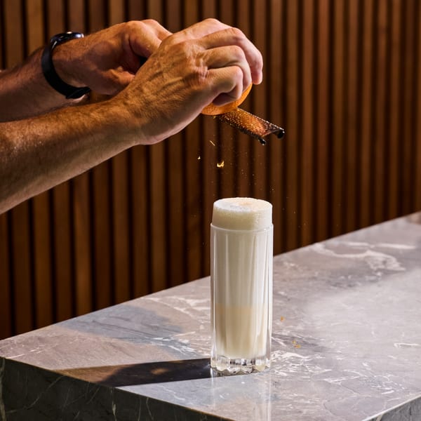 Bartender grating orange zest over a tall, foamy white cocktail on a marble bar top.