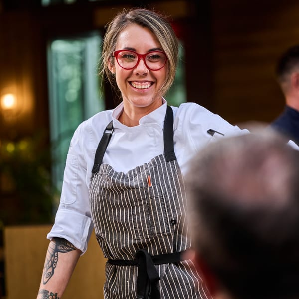 Smiling female chef with red glasses and tattooed arm wearing a striped apron at Broma Restaurant.
