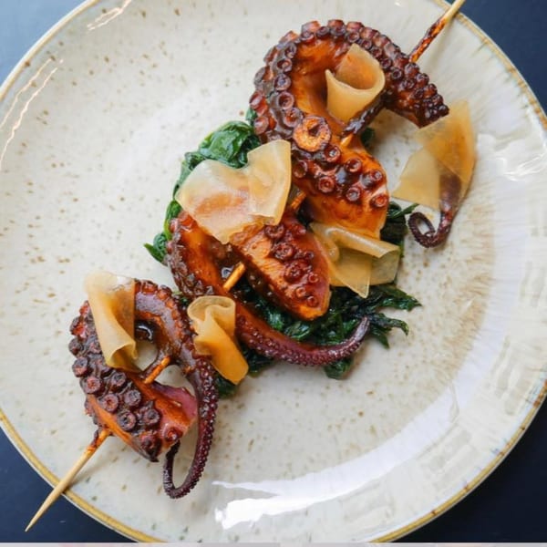 Grilled octopus skewers with greens and thinly sliced garnishes on a speckled plate.