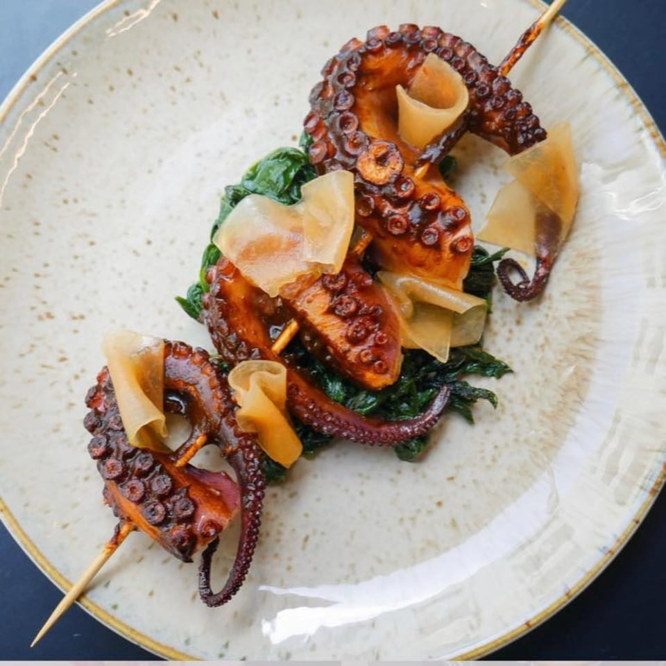 Grilled octopus skewers with greens and thinly sliced garnishes on a speckled plate.