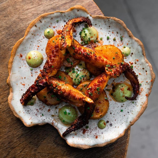 Grilled octopus with roasted potatoes, green sauce, and chives on a white and brown plate.