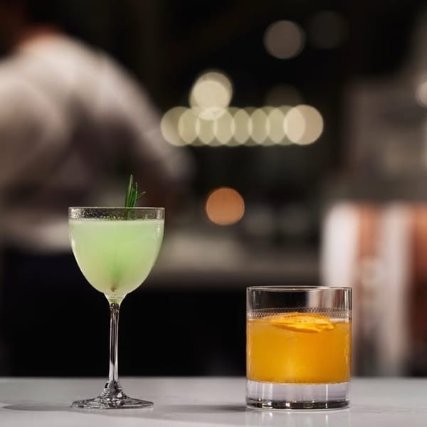 Two cocktails, one green with rosemary and one orange with citrus, on a bar at Broma Restaurant.