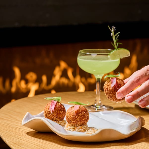 A hand places a crispy arancini ball onto a white plate, with a green cocktail and fireplace in the background.