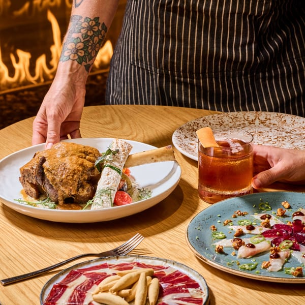 A server places a braised short rib dish on a wooden table with other appetizers and a cocktail.