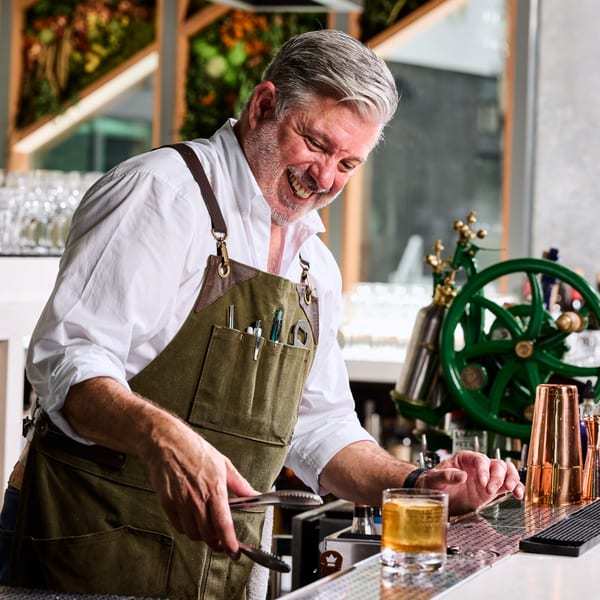 A smiling Broma Restaurant bartender in an apron prepares a drink at a bar.