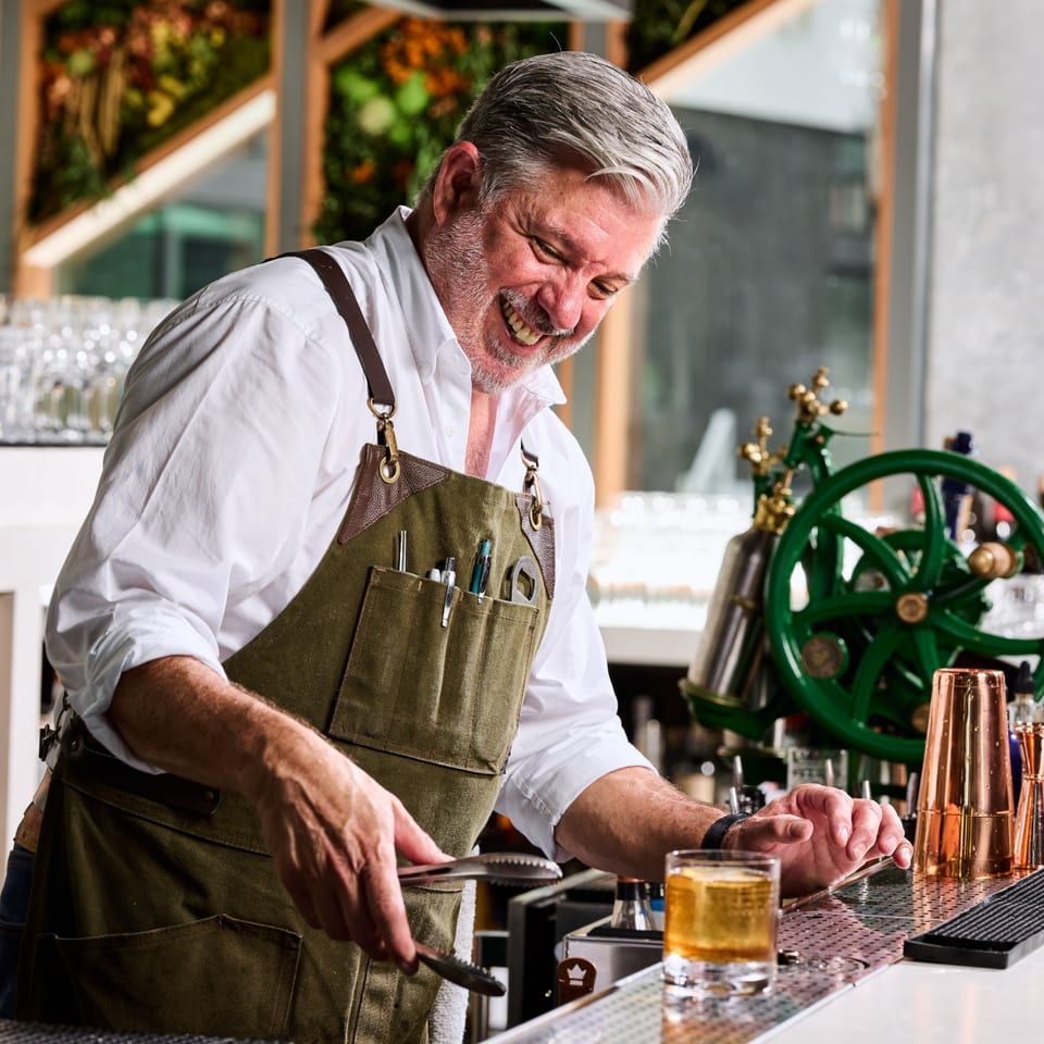 A smiling Broma Restaurant bartender in an apron prepares a drink at a bar.