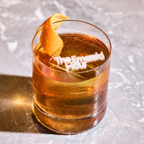 An Old Fashioned cocktail with an orange peel and a branded ice cube on a marble surface.