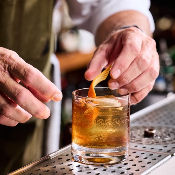 Bartender garnishing a Broma Restaurant cocktail with an orange peel on a metal bar surface.