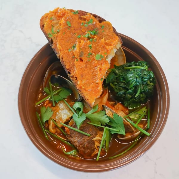 A brown bowl of Broma Restaurant's stew with clams, meat, spinach, and a slice of toast.