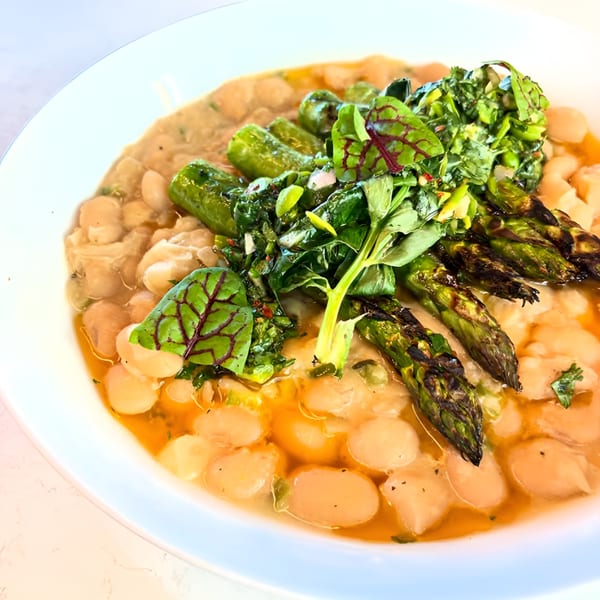A white bowl of Broma Restaurant's American cuisine, featuring beans, grilled asparagus, and fresh greens.